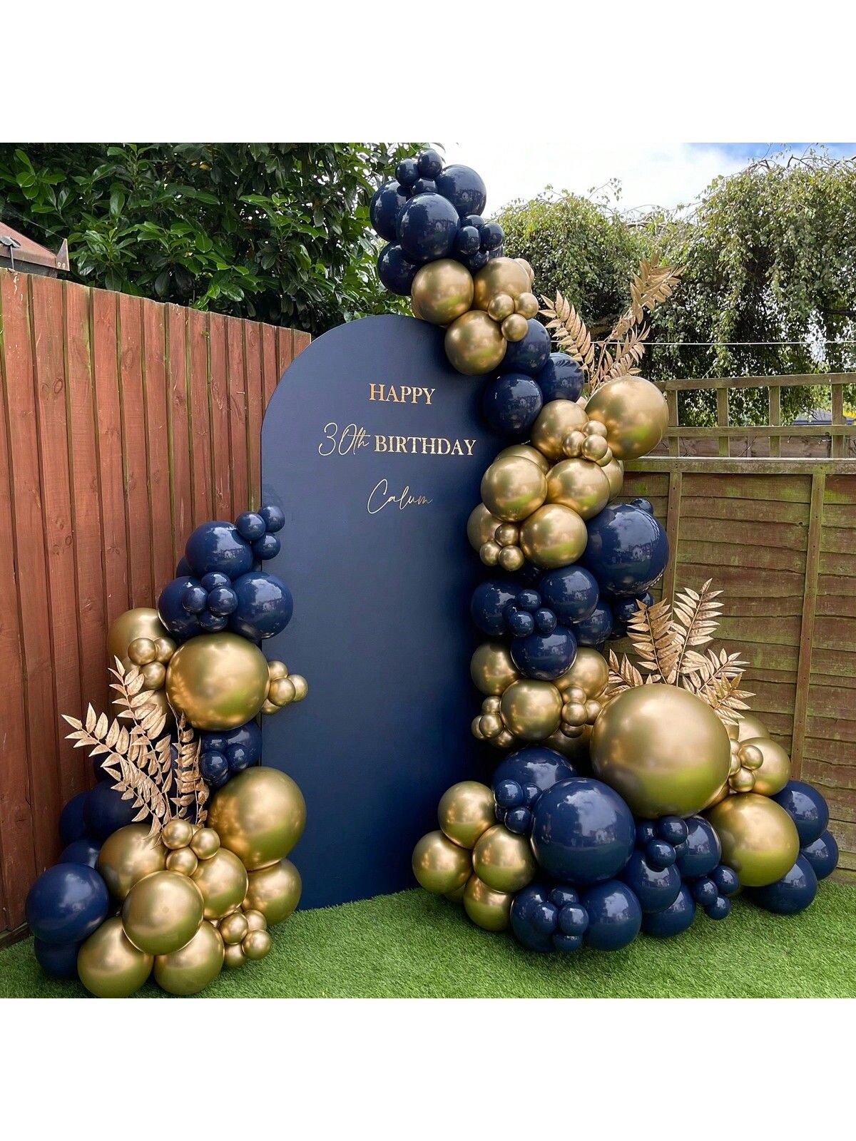 Navy arched backdrop with gold lettering framed by navy and metallic gold balloon garlands and gold palm leaves