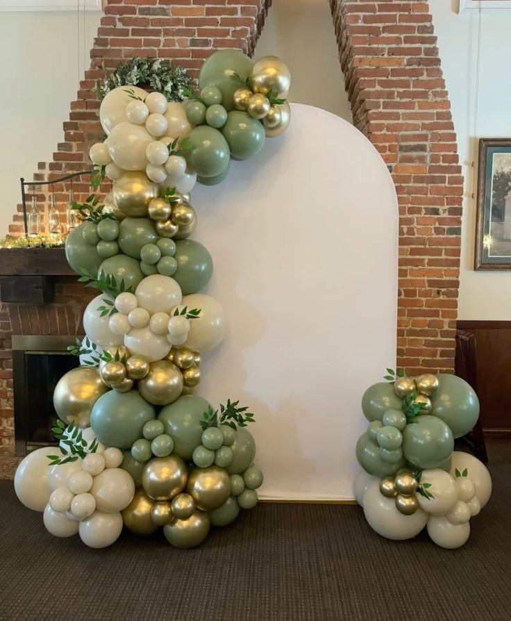 Sage green and ivory balloon garland with gold accents framing a white arched backdrop beside a brick fireplace